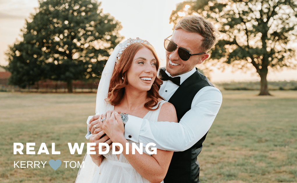 Happy married couple, very smiley with the sunset, field and trees in the background. Groom wearing sunglasses has his arms around the bride