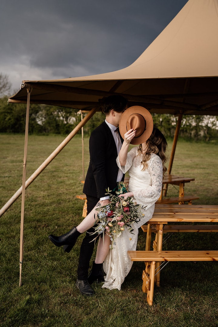 Sneaky bride and groom kiss under the little hat tipi. With Full Spring floral bouquet